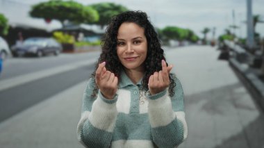 Çizgili kazaklı bir kadın hareketli bir caddede şakacı bir şekilde el kol hareketi yapıyor, açık bir şehir ortamında canlı bir fon ile para harcamayı öneriyor..