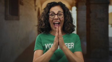 Orta yaşlı İspanyol kadın gönüllü tişörtlü, eski bir şehir caddesinde coşkuyla gülümseyen ve el kol hareketi yapan.