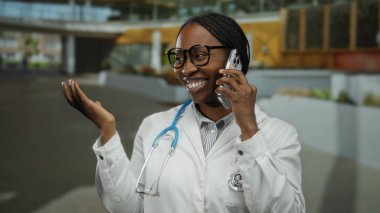 Kentsel bir bölgede, açık havada gülümseyen ve konuşan bir kadın, gözlük ve steteskop takıyor, profesyonellik ve bağlantı duygusunu iletiyor..