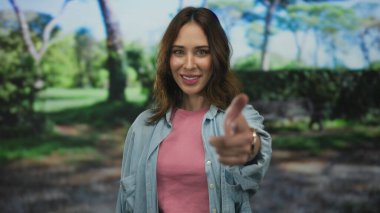 Pembe gömlekli ve kot ceketli mutlu İspanyol kadın güneşli bir parkta neşeli yeşil ağaçlar ve doğa ile çevrili.