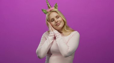 Tek boynuzlu at diademi giyen genç sarışın kadın pembe arka plana karşı komik bir uyku hareketi yapıyor.