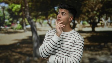 Çizgili tişörtlü, düşünceli bir genç adam güneşli bir parkta etrafı ağaçlarla çevrili, sakin bir açık hava manzarası sergiliyor..