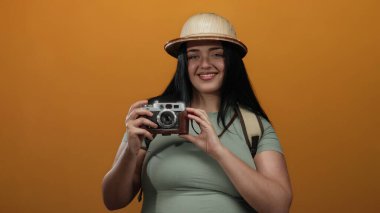 Kameralı kadın gezgin canlı turuncu arka planda poz veriyor safari şapkası takıyor güven ve macera ruhu yayıyor.
