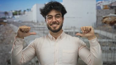 Sakallı, gözlüklü, gülümseyen ve bir inşaat alanında kendini gösteren genç bir adam, günlük kıyafetlere olan güvenini dile getiriyor..