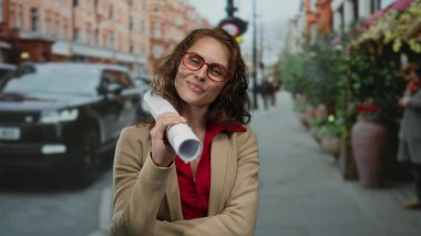 Gözlüklü, gülümseyen genç bir kadın şehir sokaklarının planlarını tutuyor. Arka planda arabaları var. Bu da iş ya da profesyonel bir ortam olduğunu gösteriyor..