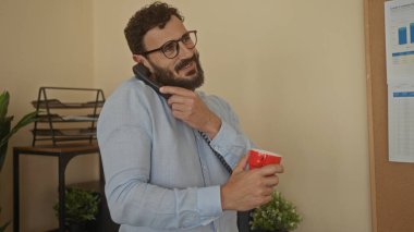 Kırmızı bardakta sakallı İspanyol bir adam ofis ortamında telefonla konuşuyor. Etrafı bitkiler ve evraklarla çevrili. Nişanlı ve profesyonel görünüyor..