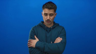 Mavi kapüşonlu genç adam, kapalı gözleri ve çapraz kolları ile izole edilmiş mavi bir arka plana karşı duruyor, düşünceli bir ruh hali içinde..