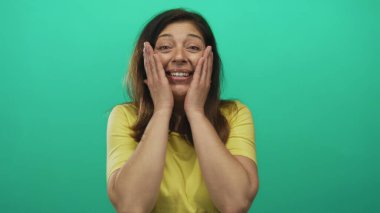 Stüdyoda iki eliyle yanaklarını tutan, ağzı açık ve gülümseyen bir kadın yeşil arka plana karşı. Sürpriz..