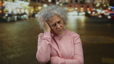 Şehir sokaklarında pembe kazaklı, gri saçlı yaşlı kadın şehir atmosferi ve bulanık geçmişiyle parlak şehir ışıkları altında düşünceli görünüyor..