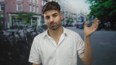 Kirli sakallı beyaz gömlekli adam, bisiklet ve tuğla binalarla kaplı şehir caddesinde sağ elini kaldırıyor..