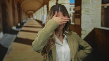 Üniversite kampüsünde beyaz bluz ve haki ceketli bir kadın parmaklarını dudaklarına uzatıyor..