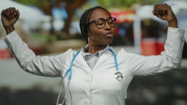 Steteskoplu beyaz önlüklü bir kadın açık havada kollarını esnetiyor. Güç, tıp mesleği, kentsel ortamdaki yetkinliği yansıtıyor..