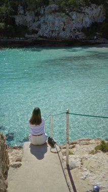 Mallorca 'daki Cala Lombars' da turkuaz su kenarında oturan kadın, güneşli ve huzurlu bir atmosfer olan Akdeniz manzarasının tadını çıkarıyor..