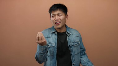 İzole edilmiş bir kahverengi duvar arkaplanına el hareketleriyle şüphecilik gösteren genç bir adam, günlük bir kot ceket giyerken belirsizlik ve tefekkür taşıyor.