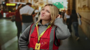 Kırmızı yelek giyen kadın şehir caddesinde, bulanık yayalar ve araçlarla çevrili, gönüllü şehir çalışmalarını gösteren hareketli bir jest yapıyor..