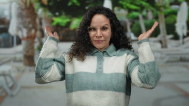 Güneşli bir sokakta gülümseyen İspanyol kadın, şakacı bir gülümsemeyle gözlerini kapatıyor ve gözlük takarak el kaldırıyor ve çizgili bir süveter giyiyor, açık havada neşe ve neşe yakalıyor..