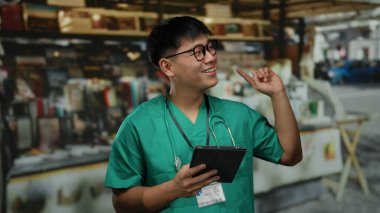 Steteskoplu yeşil önlüklü genç Çinli bir adam açık hava pazarında bir tablet tutuyor, gülümsüyor ve canlı bir şehir manzarasına bakıyor..