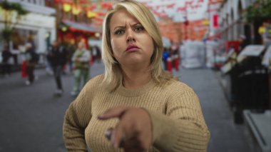 Bej renkli süveter giyen genç sarışın kadın, şehir caddesine karşı elini kalçasında gezdirerek parmak işareti yapıyor..