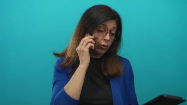 Orta yaşlı, kahverengi çerçeveli gözlüklü, mavi ceketli ve siyah bluzlu İspanyol kadın stüdyoda akıllı telefonu kulağına tutuyor..