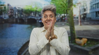 Gülümseyen bir kadın, kenetlenmiş yumruklarla gözlerini ovuşturuyor. Kanal kenarında beyaz gömlek giyiyor. Rahatlama sevinci..