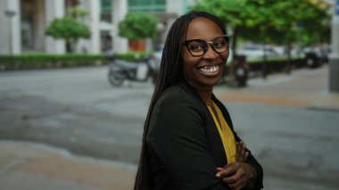 Şehir caddesinde güvenle gülümseyen bir kadın gözlük takıyor ve arka planda ağaçlar ve binalar olan sarı bir bluz giyiyor..