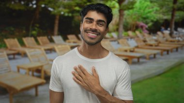 Bina terasında gülümseyen adam yüzü beyaz gömlek ve tahta şezlonglu küpeler ve görünür yeşillik; mutluluk.