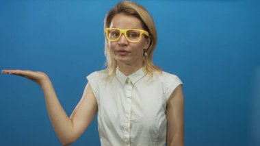 Kolsuz beyaz bluzlu sarışın kadın mavi stüdyodaki boş alana bakarken avucunu kaldırıyor..