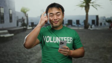 Açık havada güneşli bir günde deniz kenarındaki bir gezinti alanında elinde akıllı bir telefonla gülümseyen yeşil gönüllü tişörtü giyen genç bir adam..