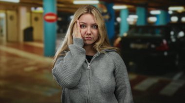 Sarı saçlı, gri kazaklı bir kadın kapalı bir otoparkta duruyor, yüzünde bir elle, arabalarla sarılmış bir şekilde..