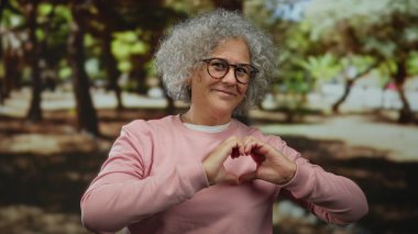 Gri saçlı yaşlı bir kadın elleri ile bir kalp şekli oluşturur sakin bir açık hava parkı ortamında dikilirken, yemyeşil ve güneşli ortamda neşe ve şefkat gösterir..