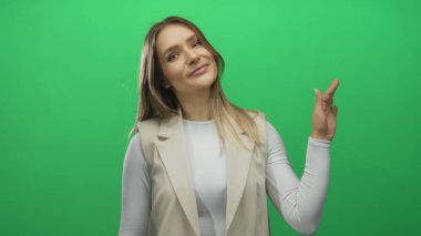 Yeşil arka plan duvarında gülümseyen bir kadın, beyaz bir gömlek ve bej bir yelek giyiyor, elini sallıyor, neşeli bir ifadeyle gündelik tarzını sergiliyor..