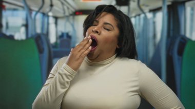 Otobüste esneyen bir kadın, kentsel toplu taşıma ortamında yorgunluğunu ifade eden, gerçek hayatta işe gidip gelme sahnesinde çeşitliliği ve duyguyu ön plana çıkaran bir kadın..