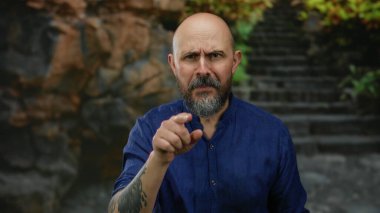 Mavi tişörtlü kel bir adam kayalık bir arka planı olan açık şehir ortamında ciddi bir şekilde el kol hareketi yapar. Yoğun ifadesini ve çekici tavırlarını yakalar..