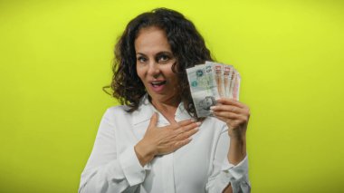 Orta yaşlı bir kadın, izole edilmiş sarı bir zemin üzerinde İngiliz sterlini banknotları tutuyor. Neşeli ve neşeli tavırlarından tatmin olduğunu ifade ediyor..