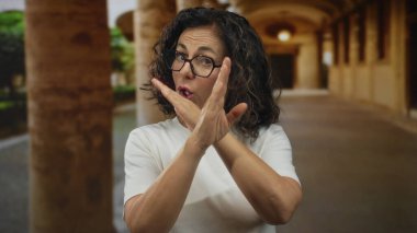 Orta yaşlı kıvırcık saçlı ve gözlüklü İspanyol kadın, eski kasaba sokaklarında reddedilme jesti olarak kollarını çaprazlıyor..