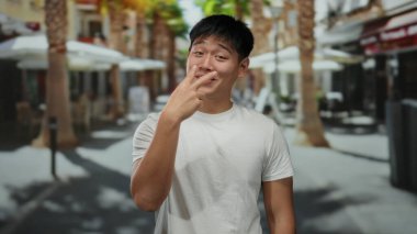 Beyaz gömlekli genç adam, güneşli bir şehir sokağında palmiye ağaçlarıyla eğlenceli bir jest yapıyor, açık hava kentsel manzarasını çekiyor..