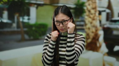 Gözlüklü, neşeli bir boks pozu veren, arka planı bulanık, çizgili gömlekli bir kadın..