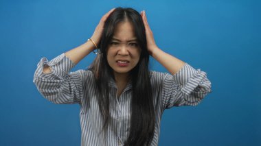 Uzun saçlı genç Çinli kadın, izole edilmiş mavi arka plan duvarına karşı kızgın bir yüz ifadesi sergiliyor, çizgili bir gömlek giyiyor ve dik dik bakıyor..