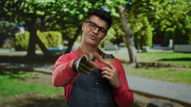 Kırmızı kazaklı genç adam yeşil bir parkta kahve fincanı tutuyor güneşli bir günde arka planda ağaçlar ve çimlerle gülümsüyor..