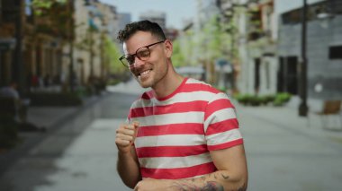 Kenar mahallede kendine güvenen genç bir adam kırmızı ve beyaz çizgili bir gömlek giyerken iki parmağını kaldırıyor..
