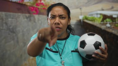 Deniz mavisi üniformalı kadın hakem iple futbol topunu kolunun altında tutuyor ve parmağıyla sokağı işaret ediyor..