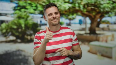 Genç adam gülümsüyor ve güneşli bir günde çizgili bir gömlek giyerken şehir ortamında başparmağıyla işaret ediyor.