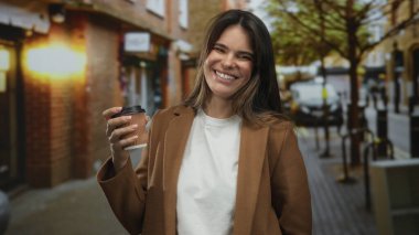Şehir merkezindeki bir sokakta gülümseyen ve kahve tutan genç İspanyol kadın kalabalık bir kentsel alanda canlı ve neşeli bir atmosfer yakalıyor..