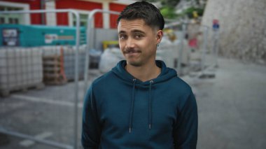 İspanyol asıllı genç adam, arka planda kentsel gelişimi vurgulayan endüstriyel ekipman ve eskrim içeren bir kapüşonlu kazak giyerek açık bir inşaat sahasında güvenle duruyor..