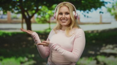 Yeşil ağaçlarla çevrili bir parkta kulaklık takan genç bir kadın güneşli bir günde müzik ve doğanın tadını çıkarıyor, rahat ve mutlu bir şekilde gülümsüyor..