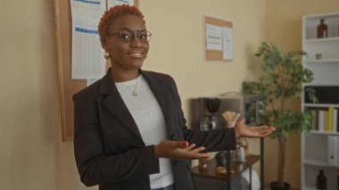 Kahverengi ceketli kadın, bina lobisinde el hareketi ve gözlükleriyle sunum yapıyor..