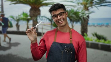 Gözlüklü genç adam elinde makasla, önlükle gülümsüyor, deniz kenarında palmiye ağaçlarıyla dikiliyor, neşeli bir berber teması yaratıyor..