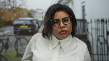 Şehir caddesinde gözlük takan bir kadın derin derin düşünüyor. Arkasına park etmiş bir araba, açık havada şehir yaşamını gözler önüne seriyor.