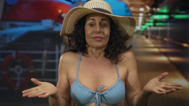 İspanyol orta yaşlı kadın bikini giyiyor ve güneş şapkası takıyor. Deniz kenarında hafif ışıklandırmanın altında dinlendirici bir anın tadını çıkarıyor..