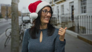 Orta yaşlı İspanyol kadın gözlüklü ve kırmızı Noel Baba şapkalı, başparmağını kaldıran ve bir şehir caddesinde gülümseyen; tatil neşesi.